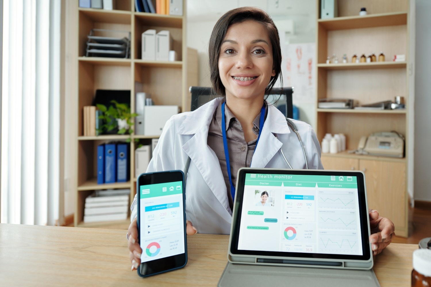 Female healthcare provider in white coat and stethoscope displaying health monitoring app on smartphone and tablet, showing patient data dashboards and medical charts in modern medical office