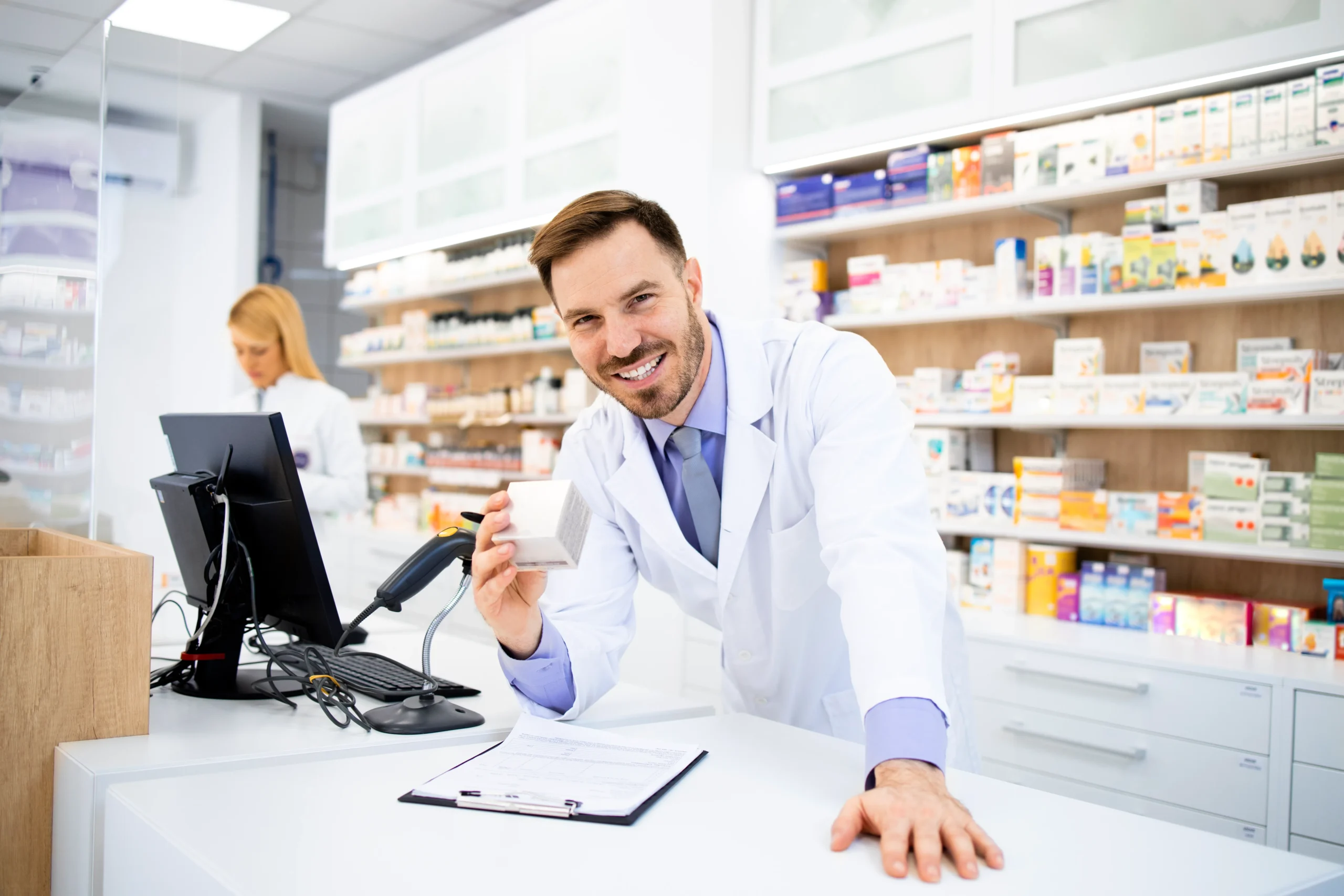 Smiling male pharmacist leaning on counter with prescription medication logged in a Pharmacy CRM system.