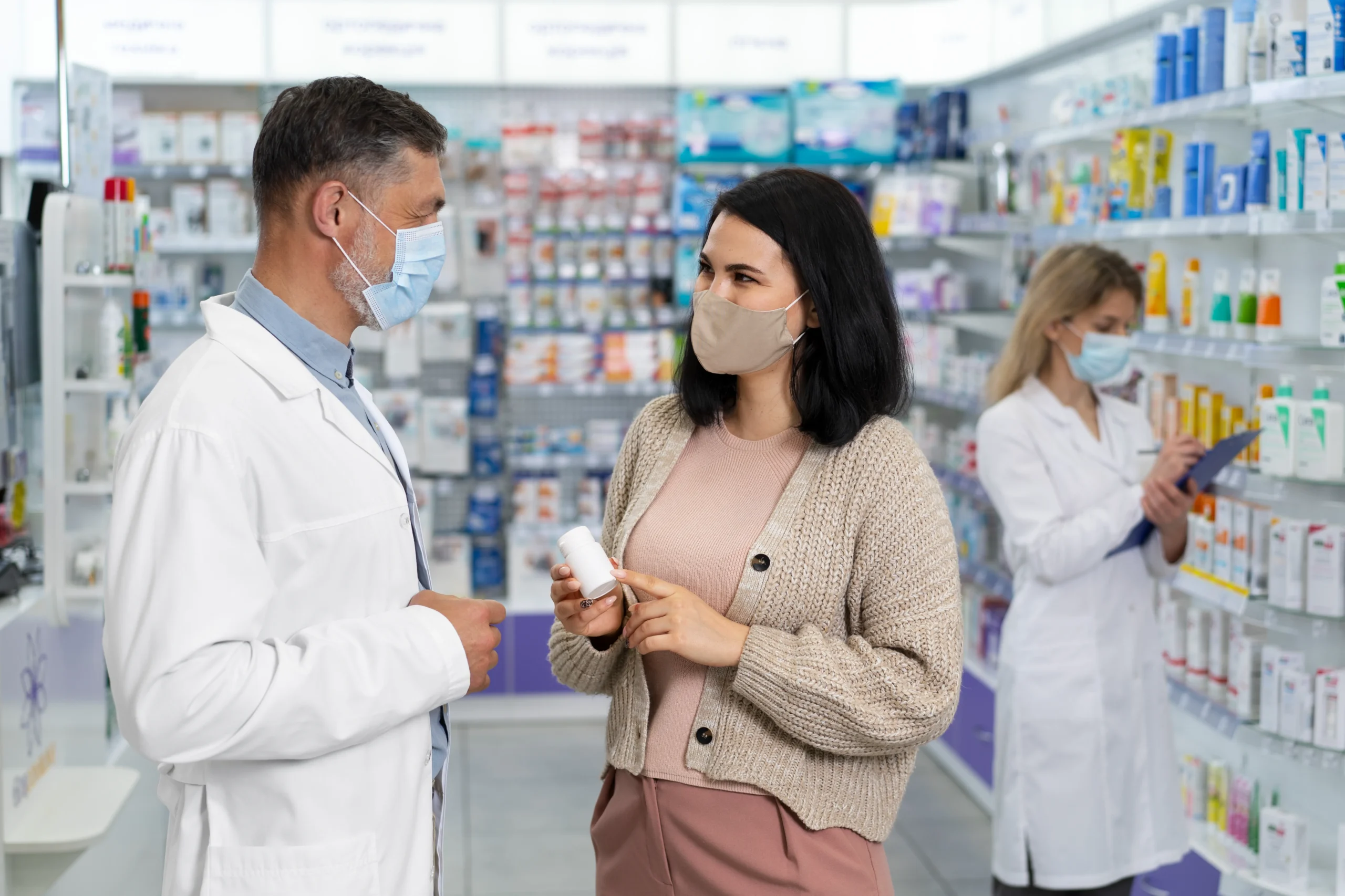 Pharmacist wearing a protective mask assisting a customer while logging visit notes into a Healthcare CRM.