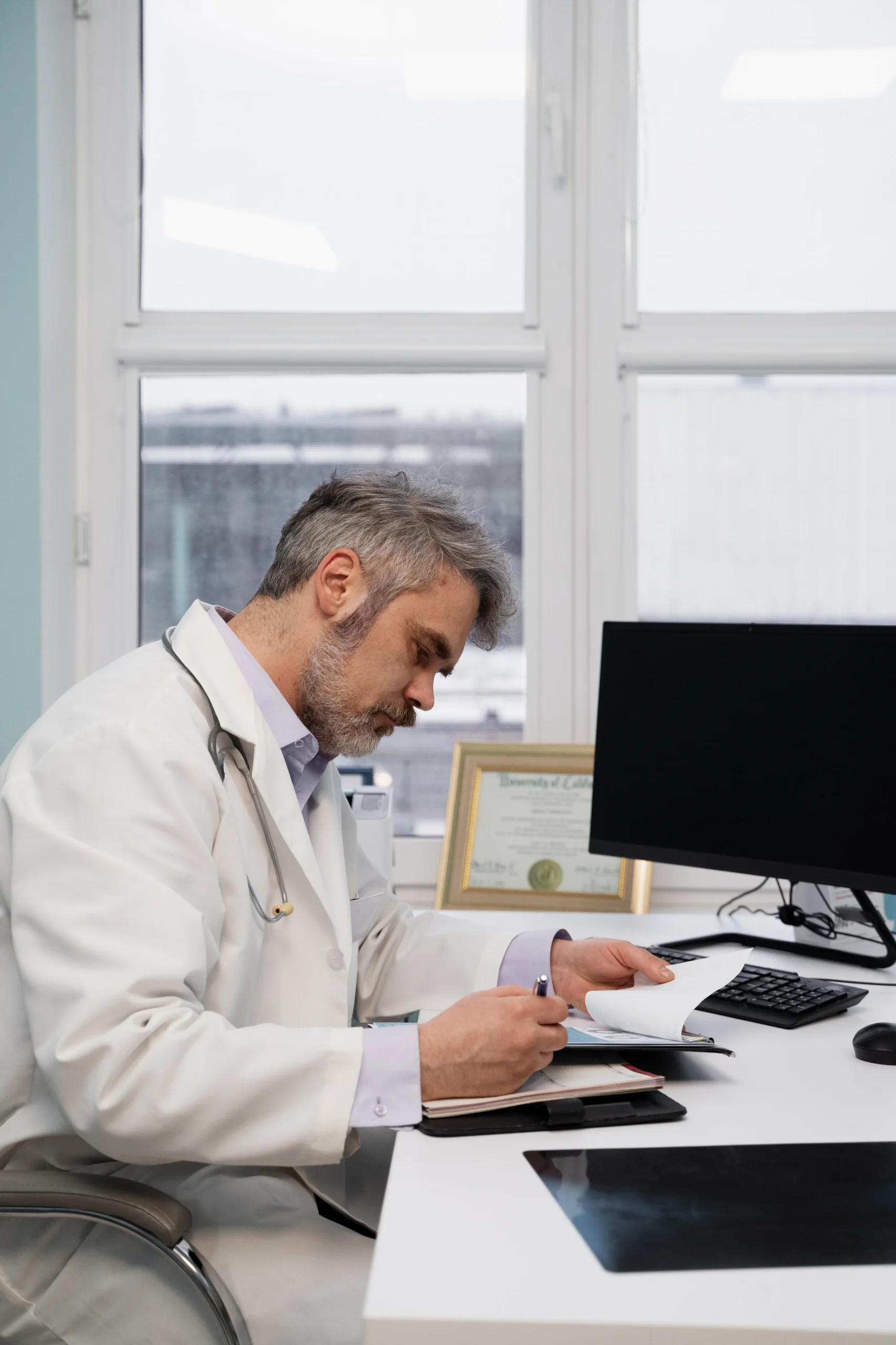 Pharmacy manager cross-referencing physical logs with independent pharmacy software on a desktop monitor.